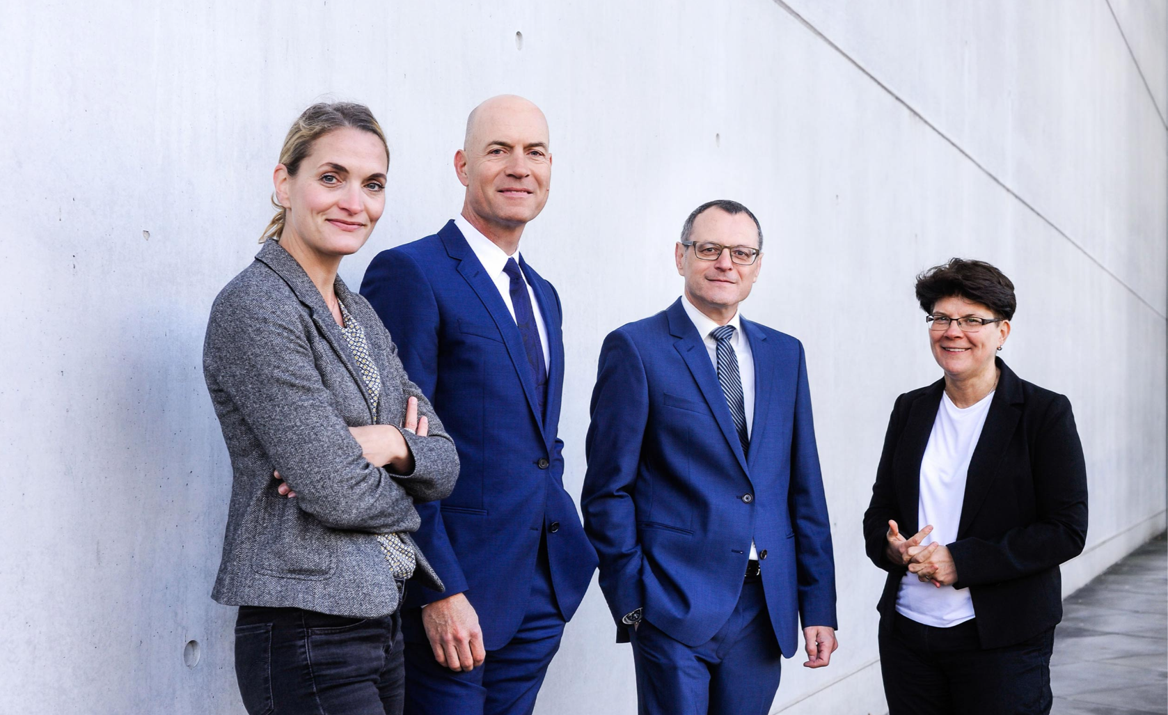 Ein Bild unserer vier Anwälte. Von links nach rechts Jutta Engels, Florian Haußleiter, Dieter Birkeneder und Karin Brandenburger. Sie stehen in Arbeitskleidung mit zuversichtlichem Lächeln vor einer grauen Mauer.