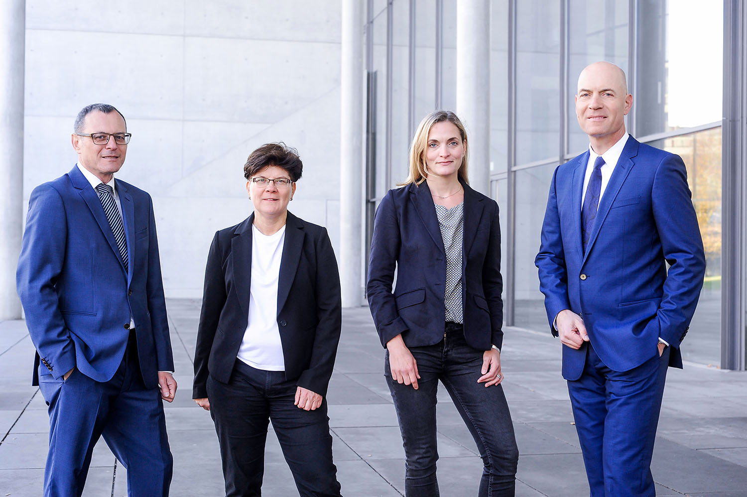 Ein Bild unserer vier Anwälte. Von links nach rechts Dieter Birkeneder, Karin Brandenburger, Jutta Engels und Florian Haußleiter. Sie stehen in Arbeitskleidung mit zuversichtlichem Lächeln vor einem grauen Gebäude mit Glasfassade..
