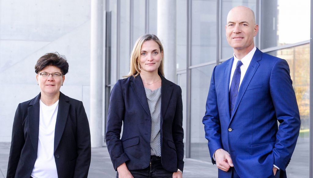Ein Bild unserer drei Anwälte. Von links nach rechts Karin Brandenburger, Jutta Engels und Florian Haußleiter. Sie stehen in Arbeitskleidung mit zuversichtlichem Lächeln vor einem grauen Gebäude mit großen Fenstern.